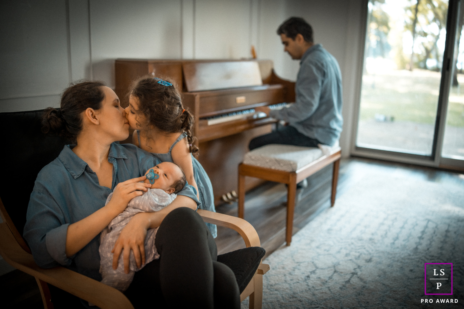 In Champaign, IL, a tender family scene unfolds as Mom kisses her daughter while holding the baby, with Dad playing softly on the piano in the background.