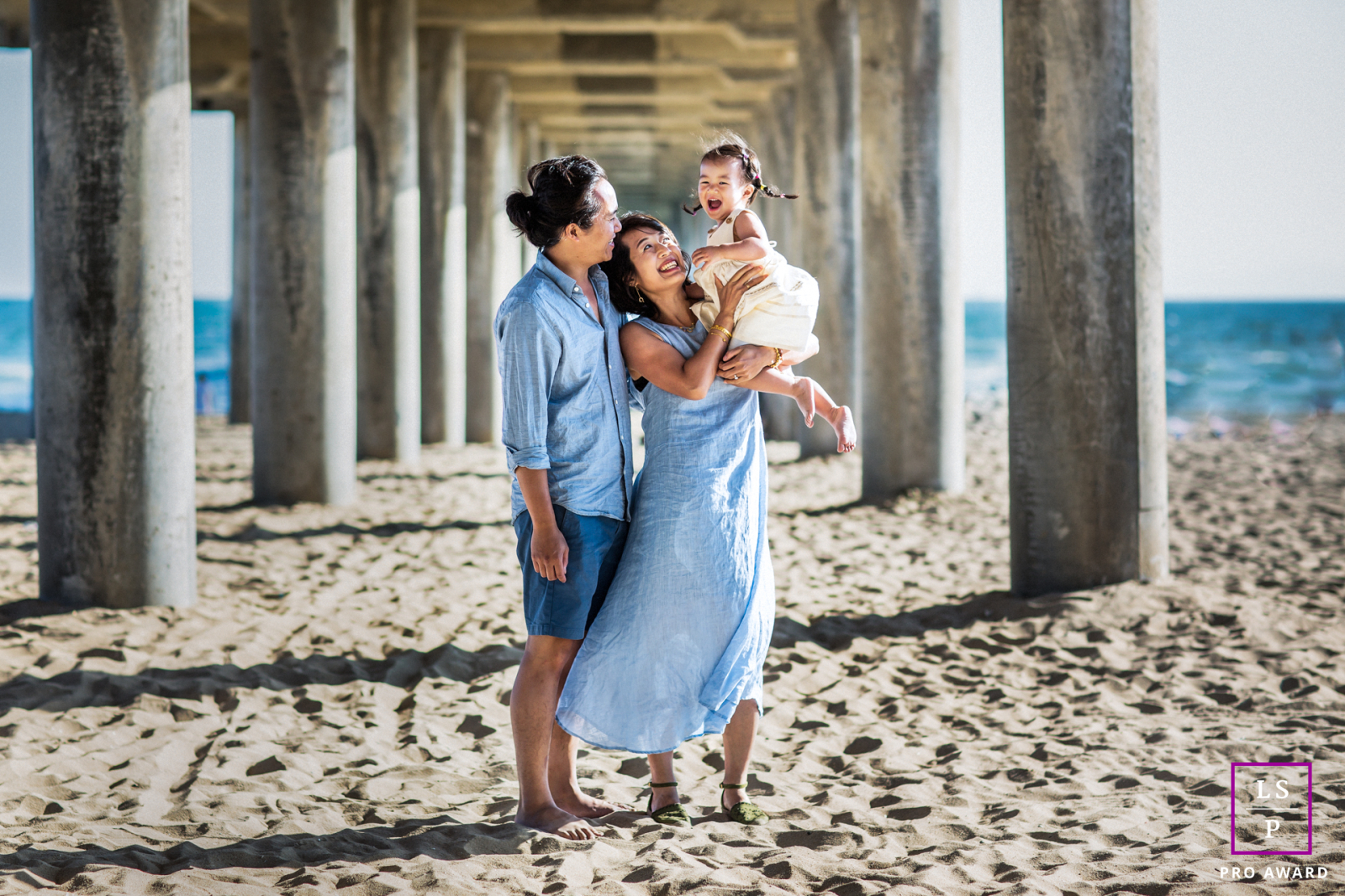 In Huntington Beach, CA, a mom and dad hold their toddler under the pier, enjoying the beach setting.