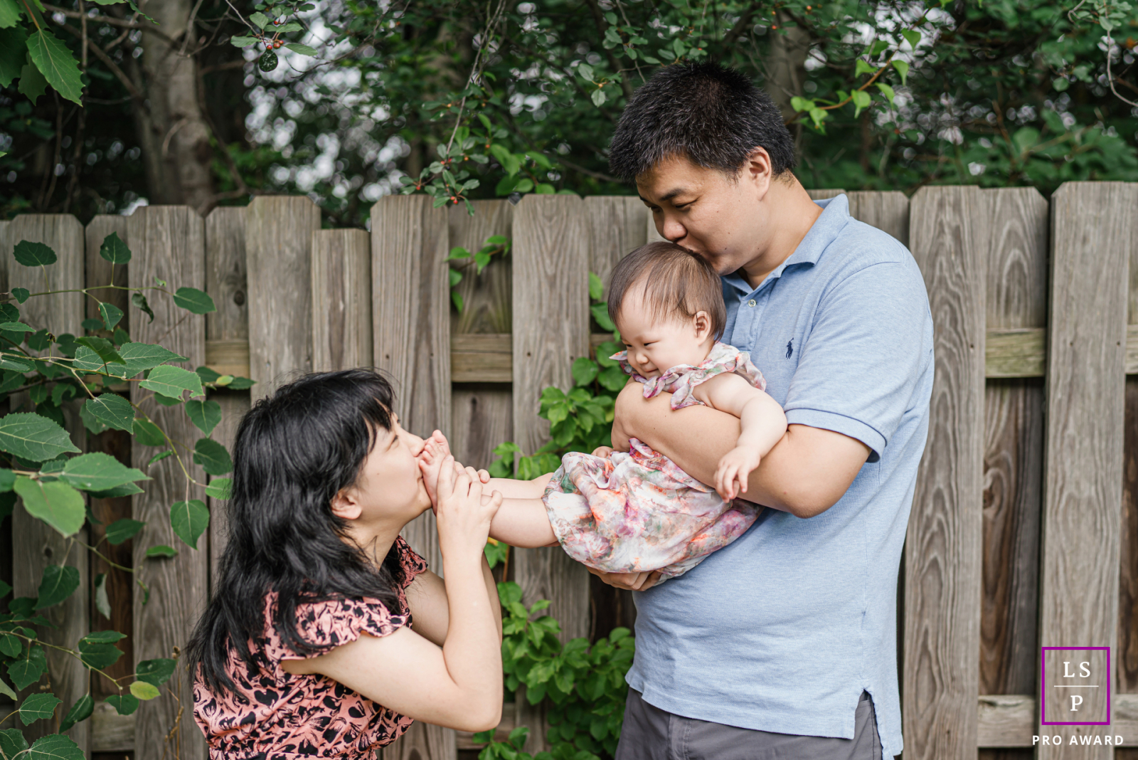 In Trenton, this baby receives kisses from both directions while resting comfortably between her parents’ affection. The playful gesture, relaxed expressions, and simple backyard setting give the photograph warmth and beautifully natural family connection