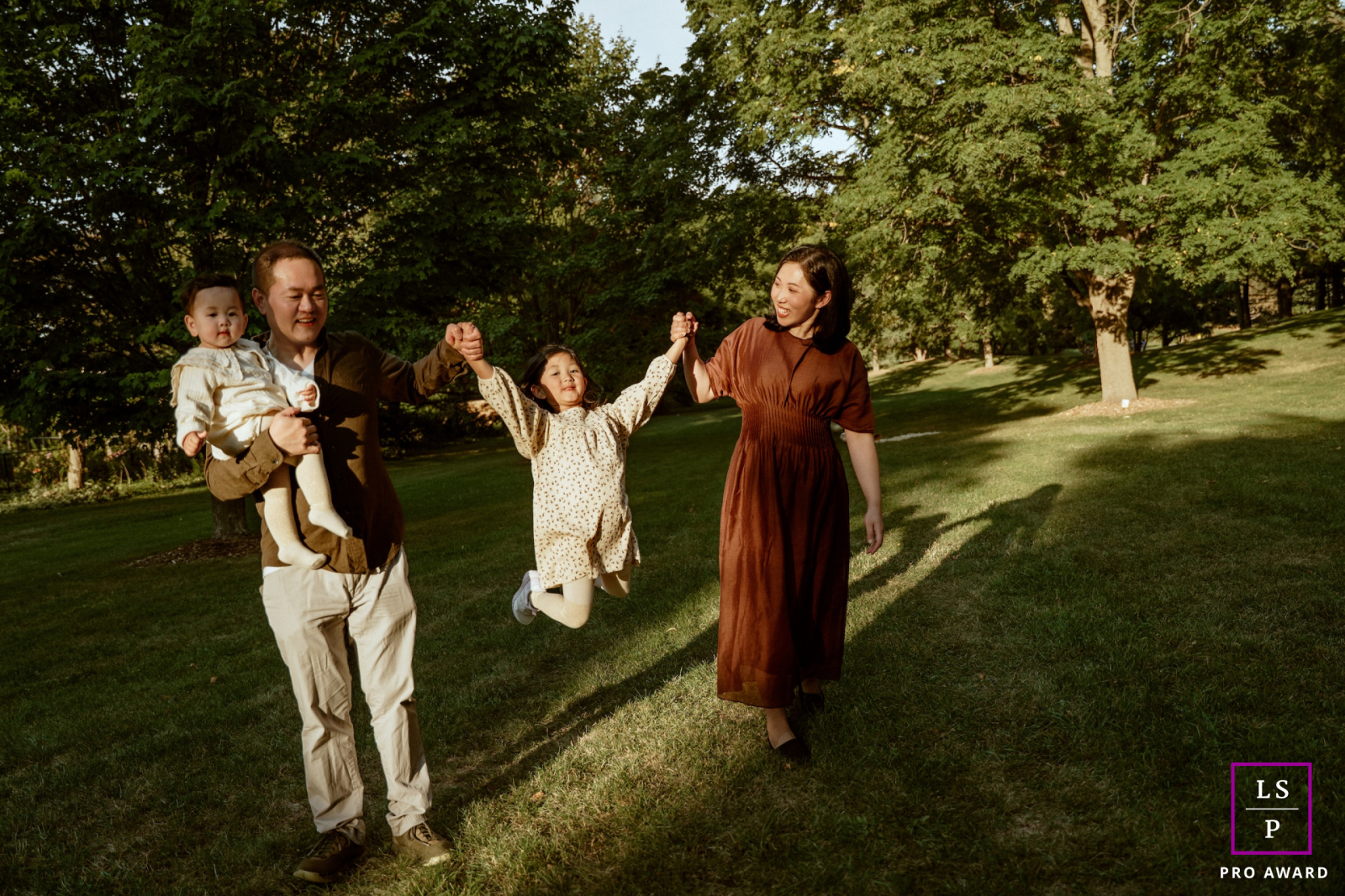 In a Toronto park, this family moves through the evening light with playful energy as one child swings joyfully between her parents. The long shadows and open green space give the photograph warmth, movement, and genuine family connection.