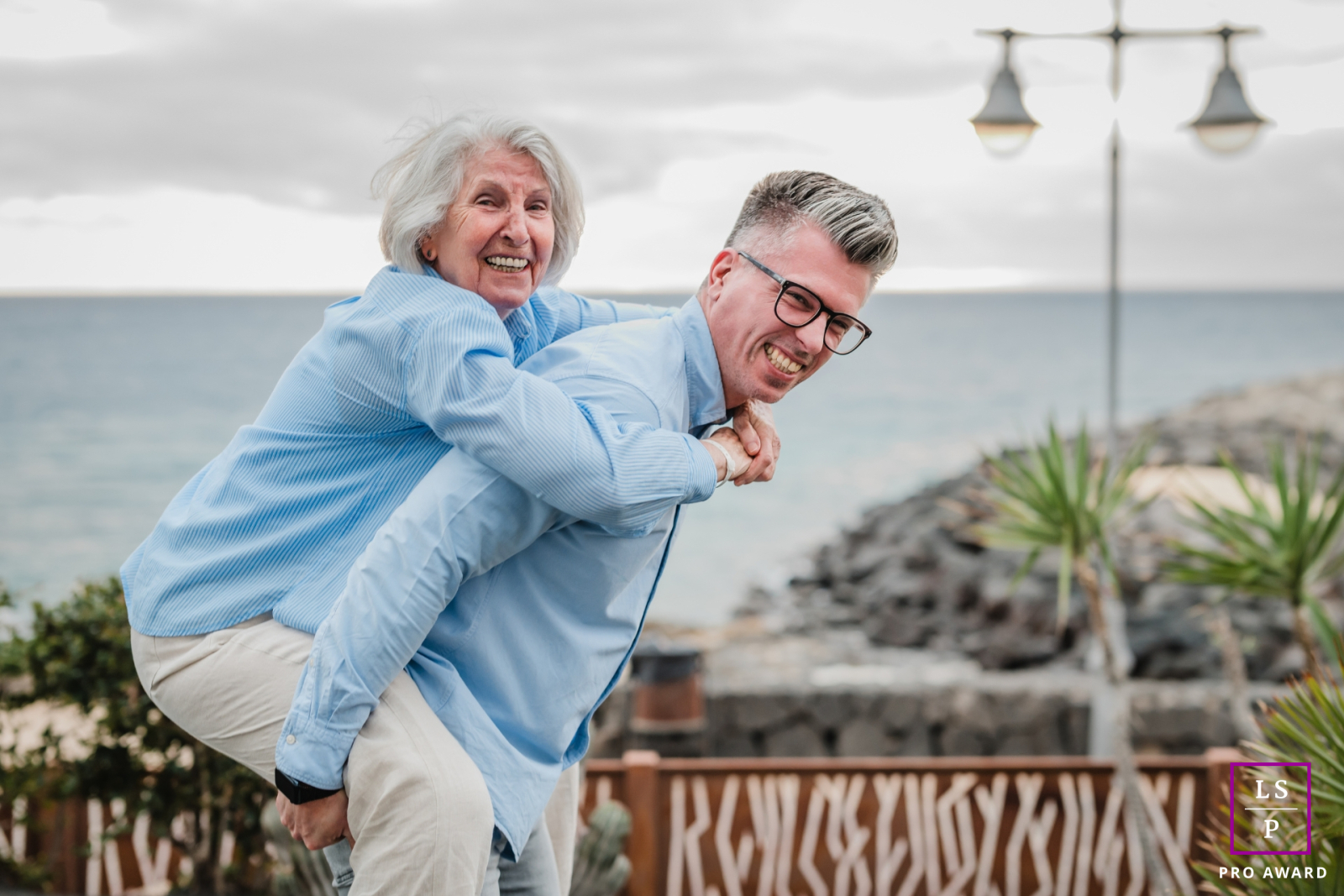 Along the coast of Lanzarote, a son lifts his mother into laughter with an unexpected piggyback ride that instantly turns playful. The seaside backdrop and matching blue tones give the photograph warmth, humor, and wonderfully youthful family energy.