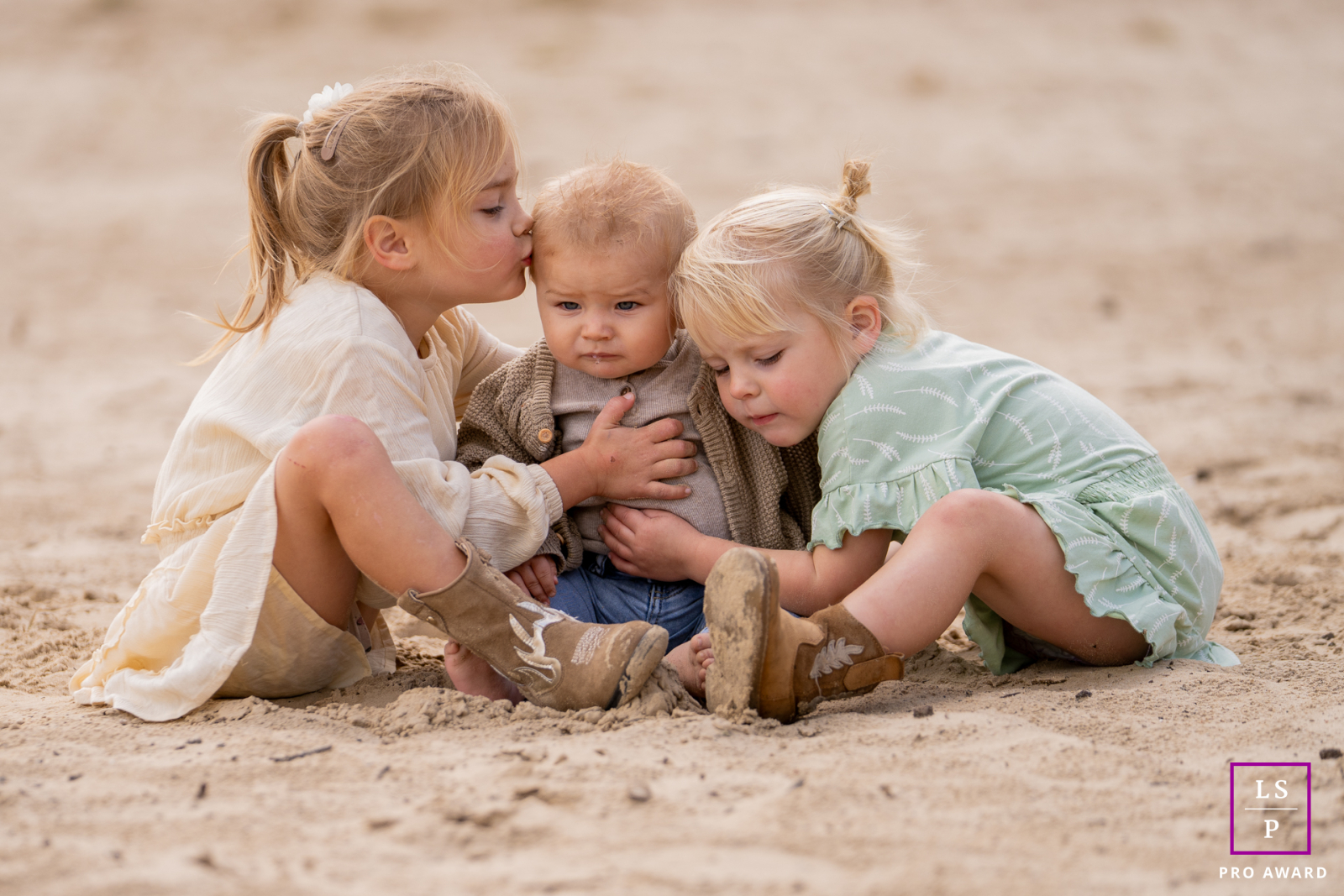 In Friesland, two sisters surround their little brother with protective affection as he quietly absorbs the attention. The soft sandy tones and close composition create a tender photograph filled with innocence, warmth, and sibling connection.