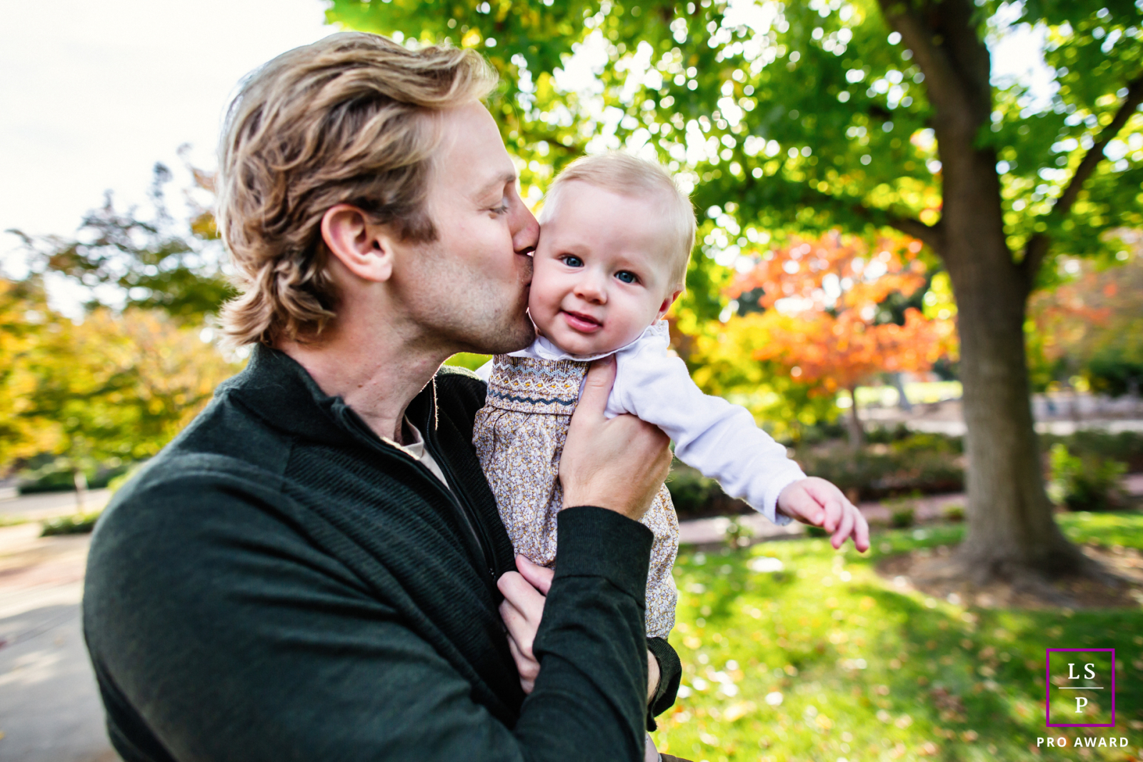 In Menlo Park, a father steals a quiet kiss while the baby looks directly toward the camera with cheerful confidence. The soft autumn color and close framing make the affection feel immediate and wonderfully sin