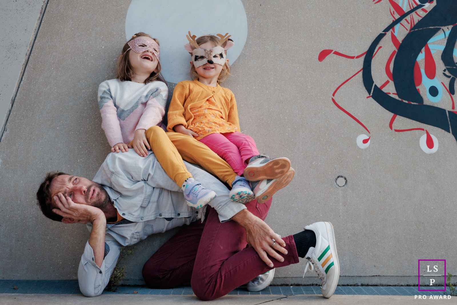 Theh exhausted Paris father becomes a human bench while the girls burst into laughter above him, completely delighted by the arrangement. The playful body language and whimsical wall art give the portrait humor, chaos, and wonderfully honest family energy