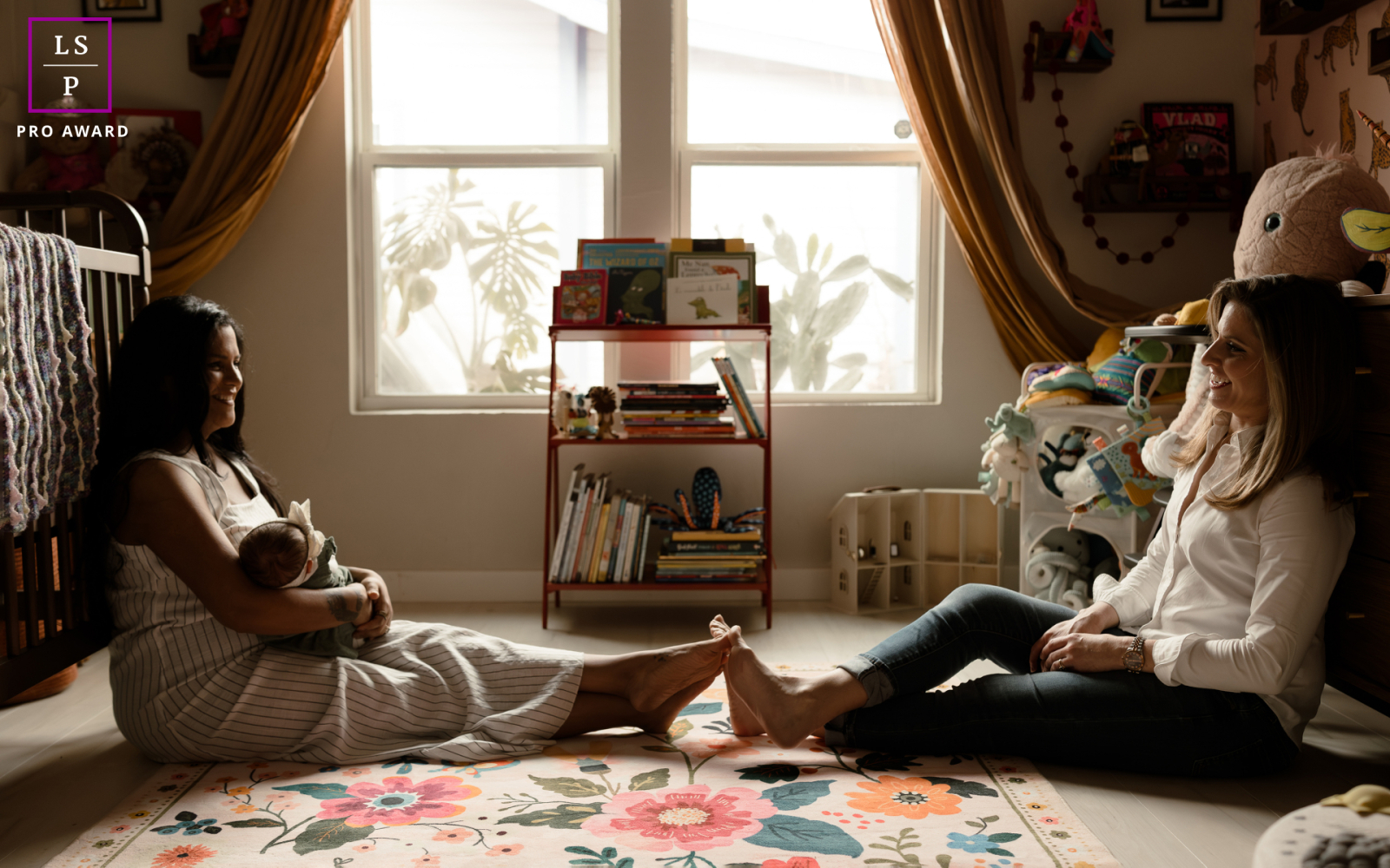 In San Antonio, Texas, two mothers sit quietly in the nursery while one cradles their newborn. The warm window light and intimate setting create a portrait filled with tenderness, calm, and the quiet joy of new parenthood.