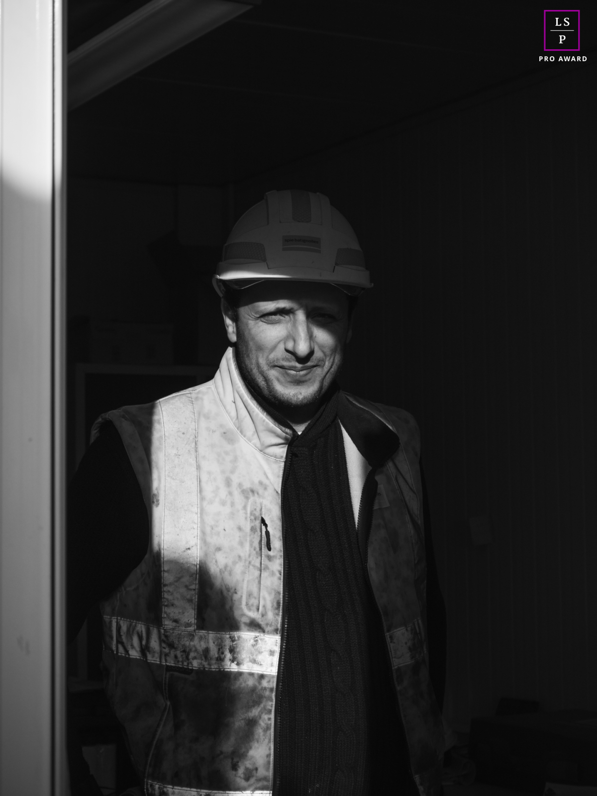 Portrait d'un ouvrier de Spie Batignolles Fondations, coiffé d'un casque de chantier, en pause sur un chantier de construction dans le Val de Marne. La lumière du soleil projette des ombres dramatiques sur son visage.