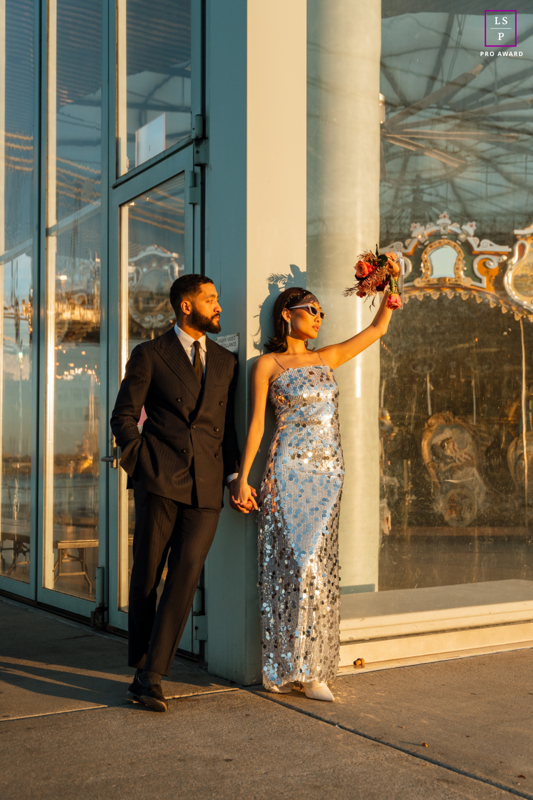 Portrait romantique en extérieur d'un couple à Brooklyn, New York. La femme tient un bouquet alors qu'ils se tiennent près d'un bâtiment en verre avec un carrousel pour enfants à l'intérieur.