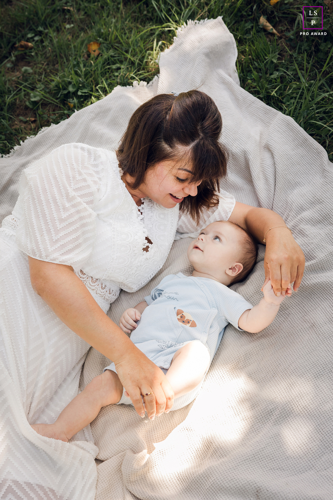 In Lyon, a mother and her baby rest together on a blanket beneath soft summer light, completely absorbed in each other’s gaze. The overhead perspective and gentle tones give the photograph tenderness, calmness, and beautifully quiet connection.