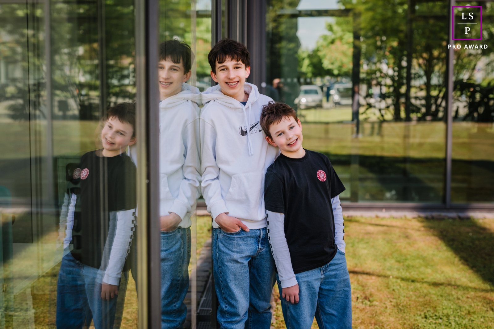 In Bonn, the brothers lean casually beside the reflective glass, creating playful doubles that quietly compete for attention. The relaxed poses and mirrored layers give the image youthful charm while adding visual complexity to keep the eye exploring.