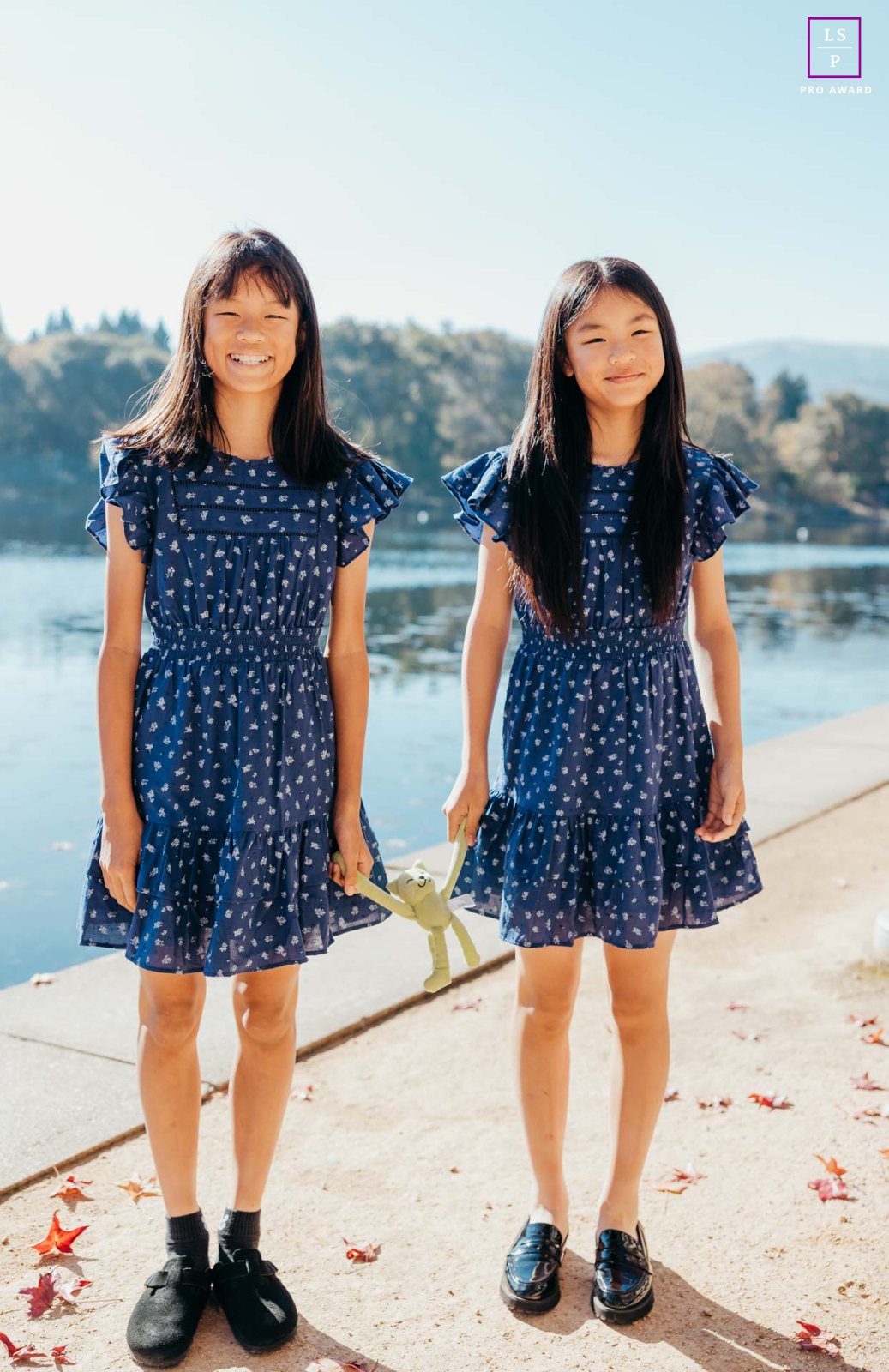 In San Ramon, California, the twins stand together in the warm fall sunlight beside the water, nearly mirroring one another in pose and expression. The soft light and matching dresses give the portrait sweetness, balance, and quiet sibling connection.