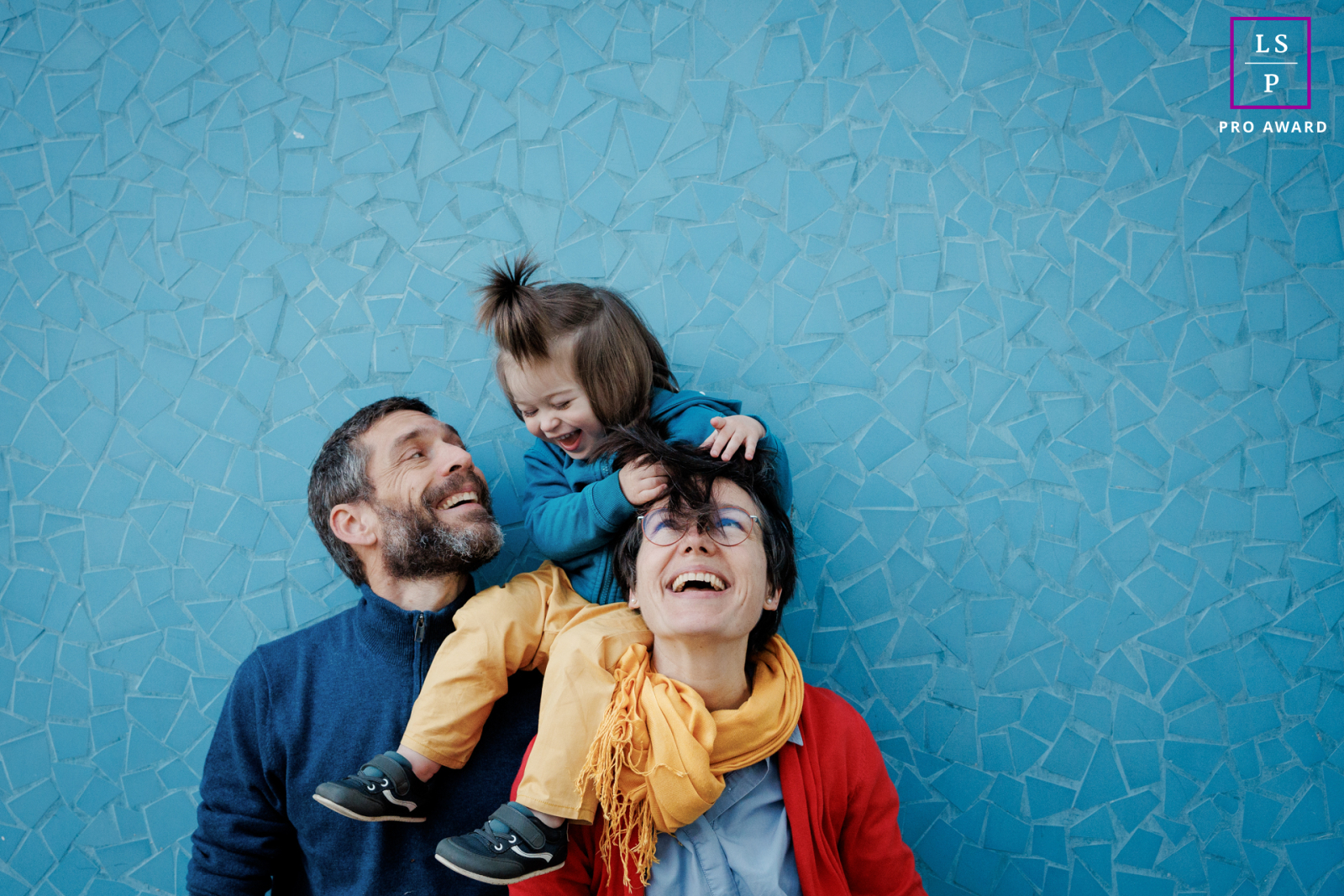 The Paris the family bursts into laughter as the child climbs onto her mother’s shoulders and grabs a handful of hair with complete delight. The bold colors and textured blue wall give the portrait playful energy, spontaneity, and joyful family chaos.