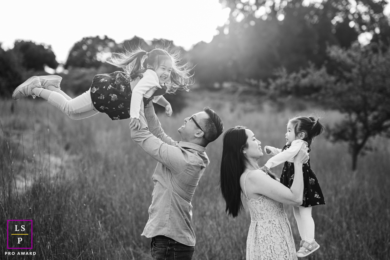 In Milpitas, California, a black and white photo captures parents lifting their two daughters, surrounded by nature and sunshine, symbolizing support and encouragement.