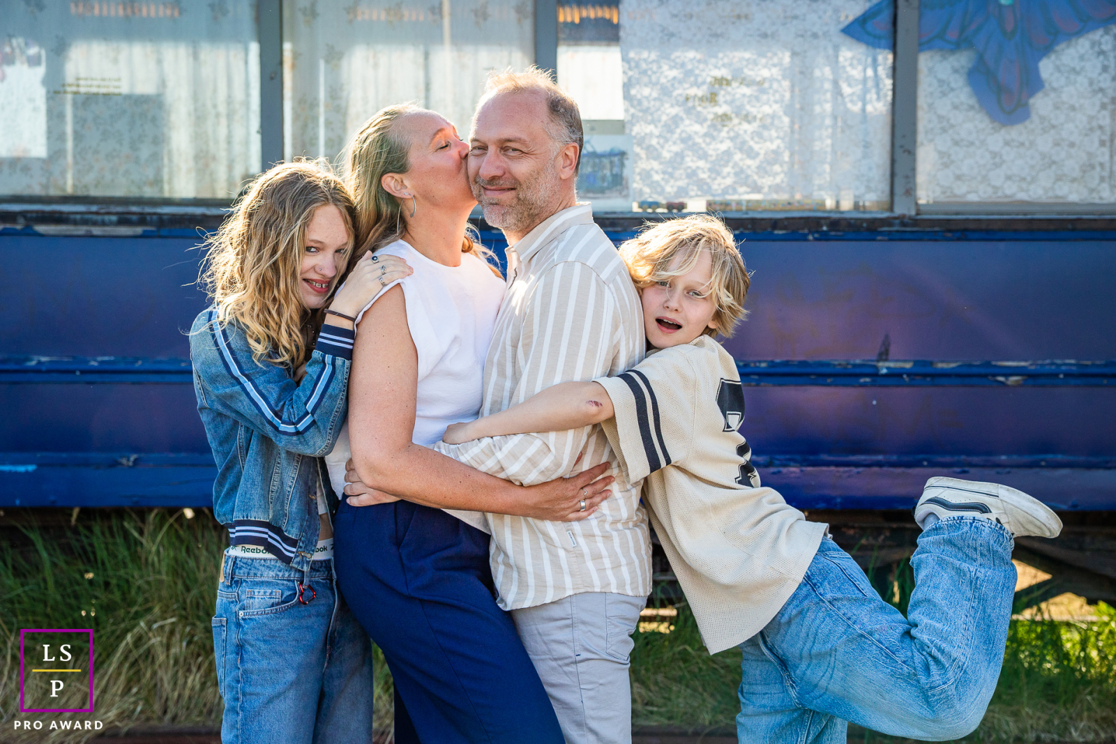 In Amsterdam, The Netherlands, mom and dad stand center facing as she kisses him. Their children on each side wrap them in tight hugs, with varied expressive faces and emotions creating a heartwarming family portrait.