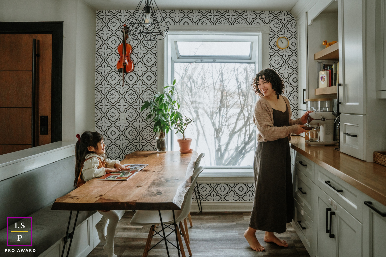 In this Toronto kitchen, a mother pauses mid task to share a warm glance with her daughter across the table. The patterned walls, soft window light, and everyday setting give the photograph comfort, rhythm, and quiet domestic beauty.