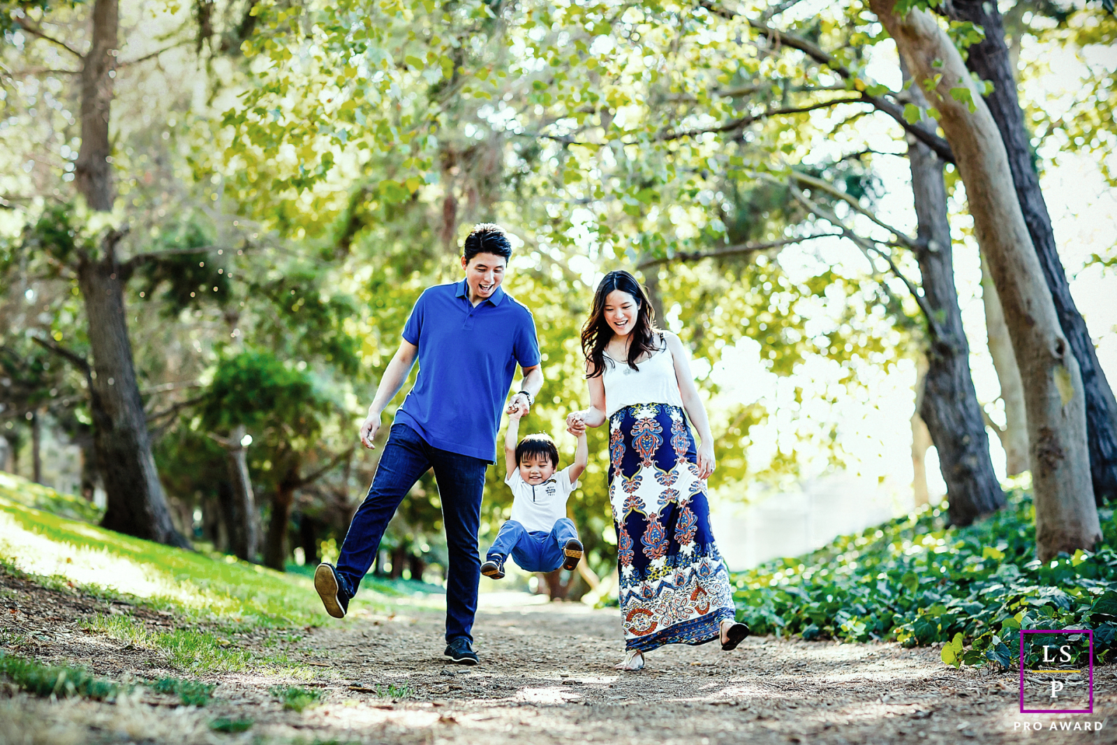 In Mountain View, CA, a family portrait captures mom and dad holding their toddler's hands, lifting the child up while walking on a tree-lined trail.