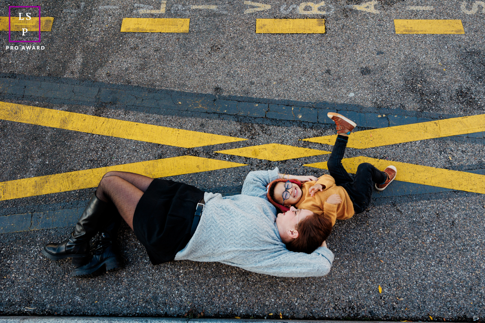 In Paris, a mother and her son playfully lie on their backs on asphalt marked with yellow traffic lines.