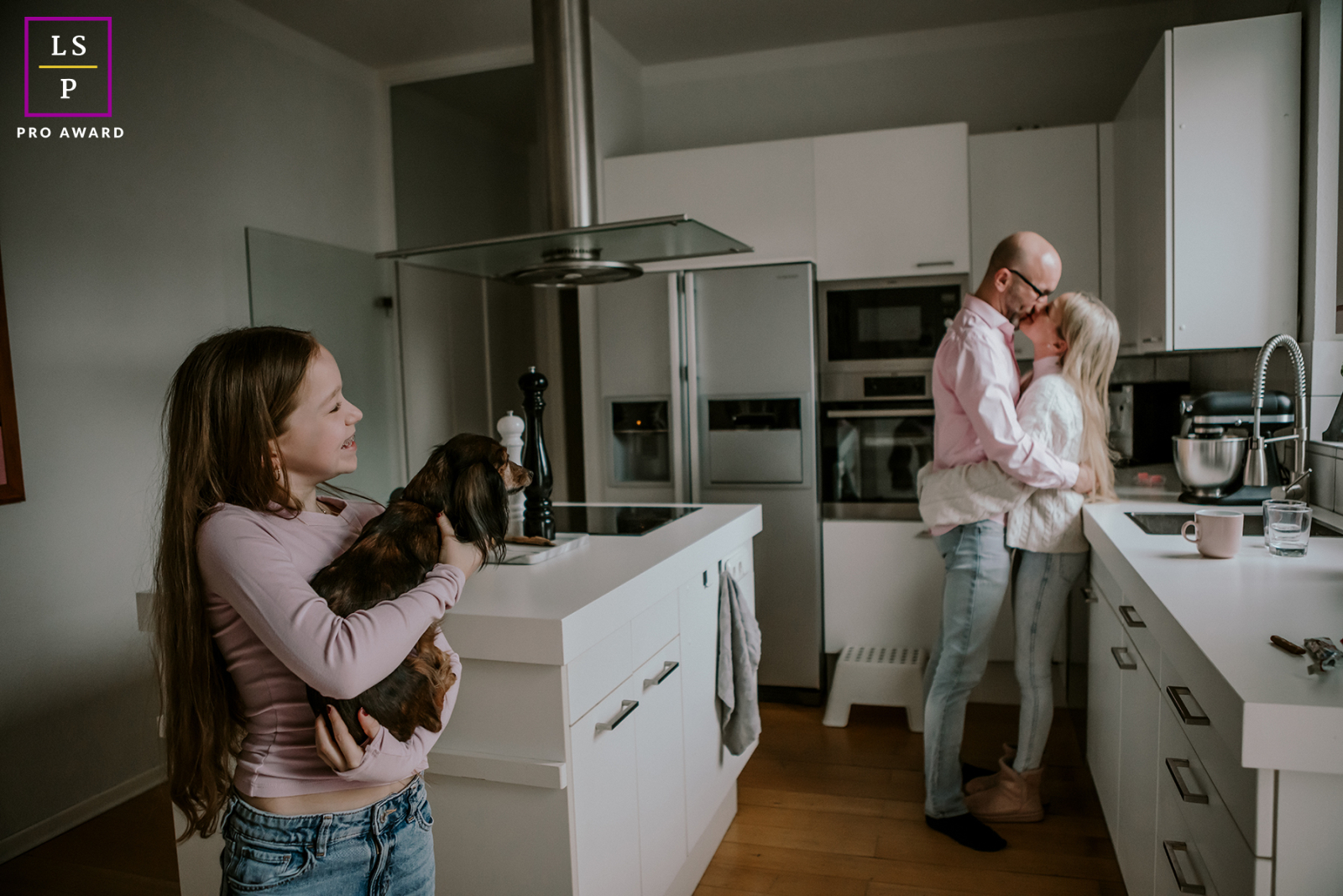 In this modern NRW kitchen, a young girl smiles while holding the family dog as her parents share a kiss behind her. The layered composition turns ordinary domestic life into something warm, playful, and beautifully connected.
