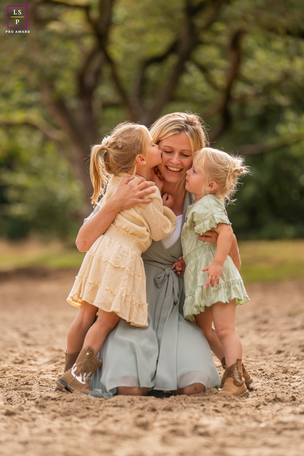 In Friesland, two little girls press close to their mother with the kind of affection that feels impossible to stage. The soft earth tones, blurred woodland backdrop, and circle of embraces turn simple cuddles into something deeply warm and genuine.