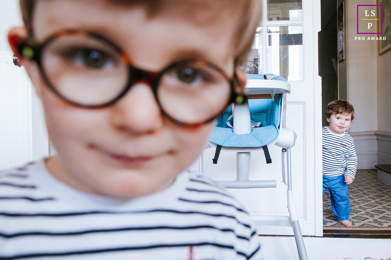 In this Mauleon home, one child dominates the foreground with oversized glasses while a sibling quietly watches from the doorway behind. The playful depth and layered composition give the photo humor, curiosity, and wonderfully authentic family energy.
