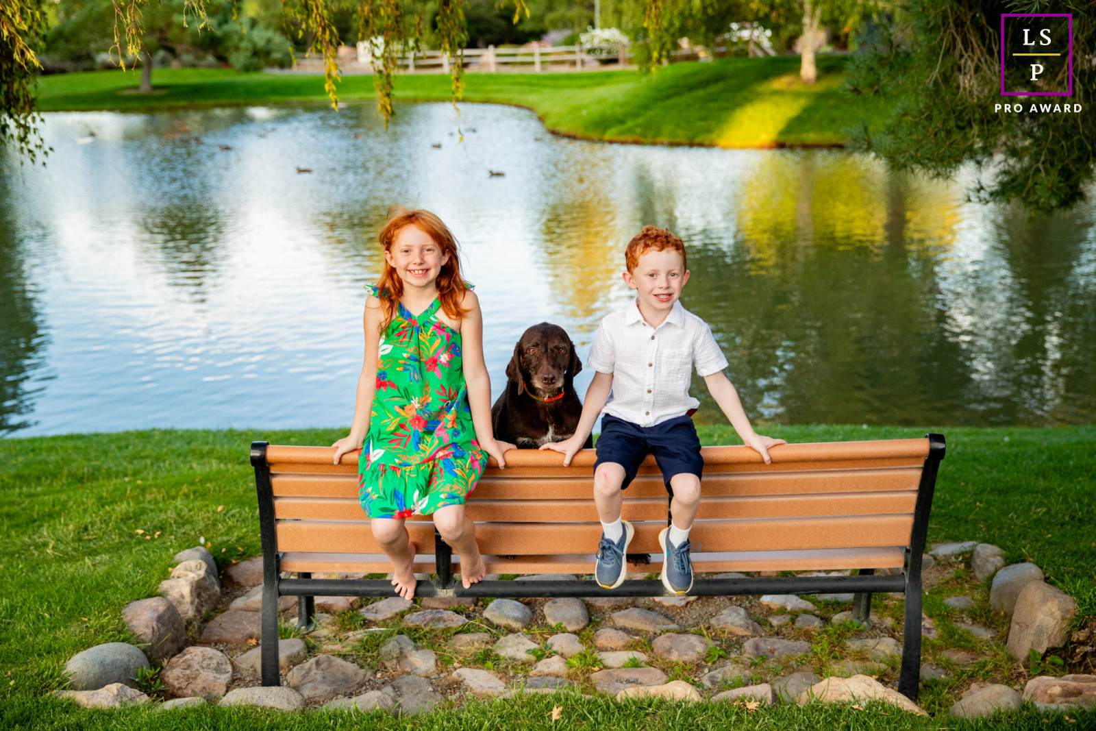 Beside the calm Reno pond, two children and their loyal dog sit proudly on a park bench like lifelong friends. The symmetry, bright color, and relaxed expressions make the photograph feel effortlessly cheerful and timeless.