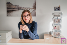 Sylvia Kaufmann Holds Coffee at Welcome Desk in Calm, Inviting Studio in Hagen, Germany Portrait At the welcome desk in Hagen, Germany, Sylvia Kaufmann holds a warm cup of coffee as light fills the studio. The soft tones, travel prints, and relaxed expression create a portrait that feels approachable, calm, and quietly inviting anyone walking in.