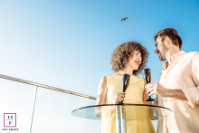 Couple toasting with champagne at a glass table, glass railing, blue sky, and a bird flying.
