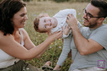 Young Child Laughs and Plays Between Seated Parents in Hauts-de-France Park A joyful family captured in a candid photo as a young child plays and laughs between their seated parents in a park in Hauts de France.