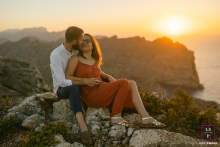On Mallorca Island, Spain, a couple sits on a cliff above the ocean, captured in the warm glow of sunset. The scene highlights their serene connection amidst the breathtaking coastal view.