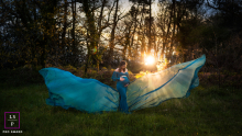 A maternity photoshoot is taking place in Saucats, France. The pregnant woman is wearing a dress, and is backlit by the sun in a forest. The dress is fanned out, resembling butterfly wings.