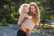 In Noord-Holland, The Netherlands, a mother with sunlit, curly hair warmly looks at the camera while her smiling daughter rides on her back. Both are rimlit by golden sunlight, radiating joy.