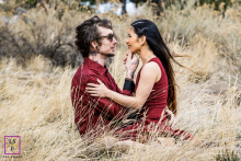 A Gentle Breeze Whispers Through Denver's Tall Grass In Denver, CO, a couple sits snuggled together in tall grass, both dressed in burgundy clothing. The wind gently blows her hair, adding a romantic, natural touch to the serene scene.