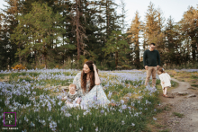 In Camas, WA, a mom lovingly sits and watches her little one playing with wildflowers, while dad and another child stand together in the softly blurred background, enjoying the peaceful scene.
