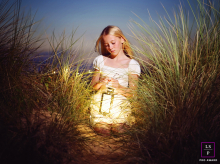 At St Annes Beach in the UK, a young girl kneels near a lantern by tall grass on sand dunes as the light fades in the evening.