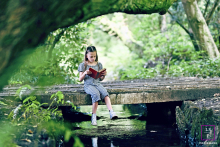 In Blackpool, UK, a young girl sits on a wooden footbridge by a stream, reading a book and appearing carefree in the tranquil setting.