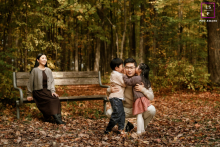 In Toronto, Ontario, a family enjoys an autumn afternoon; mom sits on a wooden bench in the background while dad hugs their two children in the foreground.