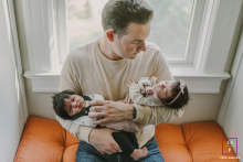 In Bergen County, New Jersey, a father sits in a bay window, holding his newborn twins in a tight embrace. The symmetrical top-down shot highlights his protective hold.