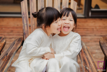 Happy identical twins in Potomac, MD, sit together on a chair, cheek-to-cheek, pressing their faces together joyfully.