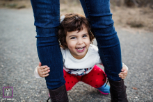 A small toddler in Cupertino, California, smiles at the camera while peeking between their parents’ spread legs in a solo portrait.