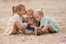 In Friesland, two sisters surround their little brother with protective affection as he quietly absorbs the attention. The soft sandy tones and close composition create a tender photograph filled with innocence, warmth, and sibling connection.