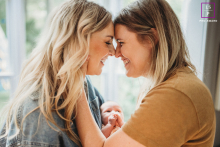 Two Mothers Smile Together While Holding Their Newborn In Boston Two mothers touch foreheads and smile warmly while holding their newborn between them during a tender family session at home in Boston.
