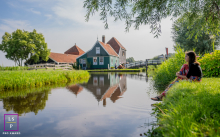 Woman Sits Beside Water Overlooking Dutch Buildings At Zaanse Schans A woman sits quietly beside the water, overlooking reflections of traditional Dutch buildings at Zaanse Schans in the Netherlands.