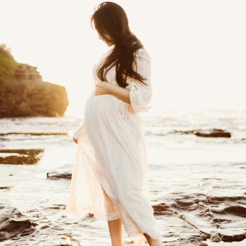 This stunning lifestyle portrait captures a serene sunrise maternity beach session in Sydney, New South Wales, with the mother beautifully alone near the water wearing a white dress
